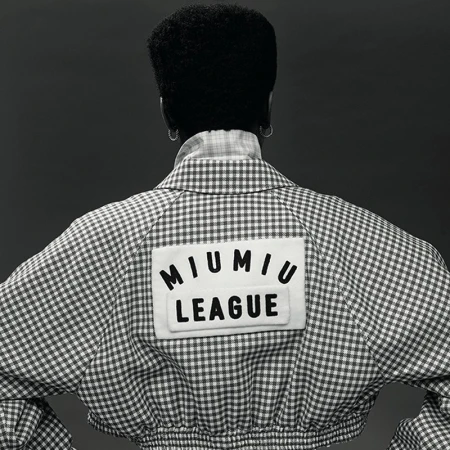 A woman with dark hair in a white and grey gingham jacket featuring a 'Miu Miu League' patch, captured in a back-facing studio shot.