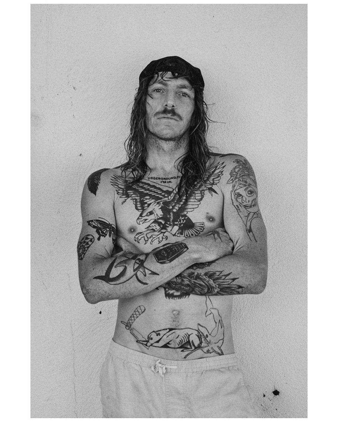 Black and white portrait of shirtless adult male with tattoos, wearing a cap, standing against a light neutral wall, arms crossed.
