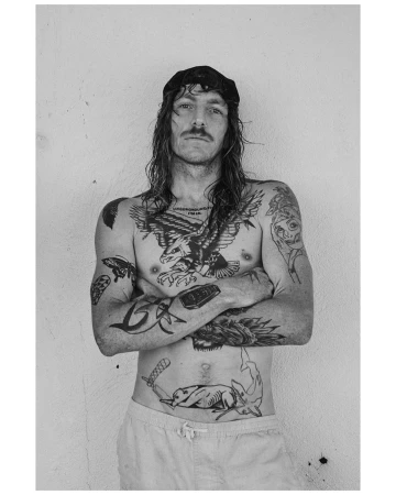 Black and white portrait of shirtless adult male with tattoos, wearing a cap, standing against a light neutral wall, arms crossed.
