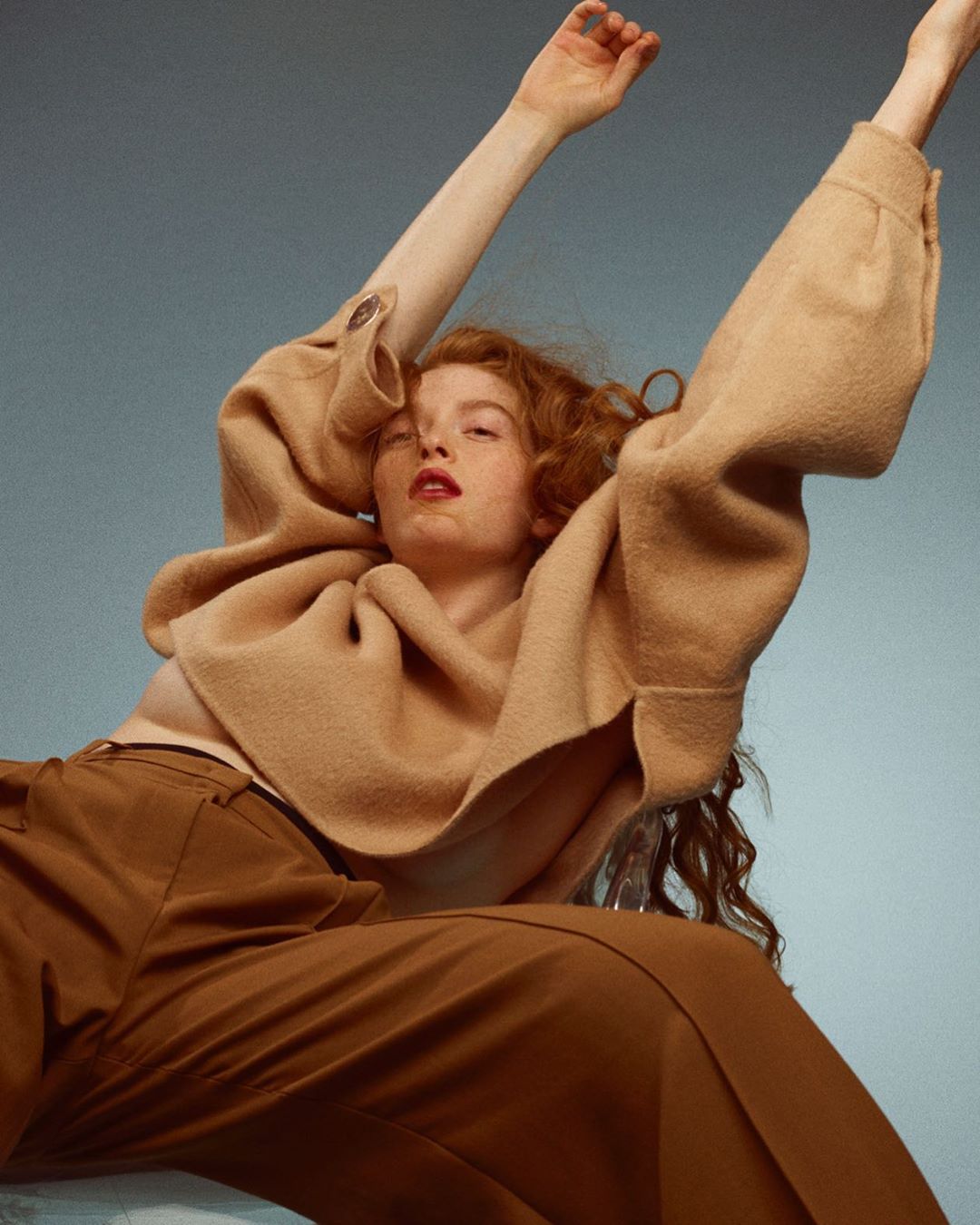 A fashion shot presents a red-haired woman in a draped tan top and brown slacks, set against a blue-grey backdrop, with arms raised.