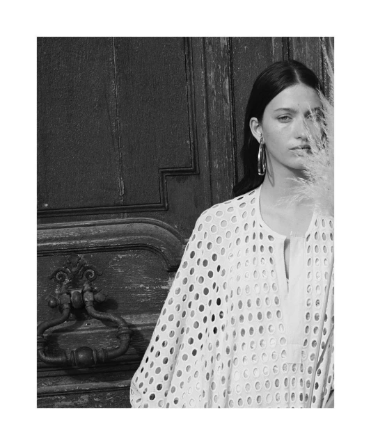 Black and white portrait of a woman in a perforated top standing in front of an ornate wooden door, framed by a sprig of pampas grass.