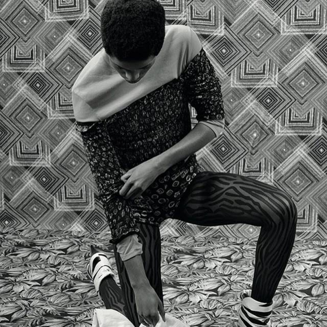Monochrome fashion shot of a person kneeling on a patterned surface, wearing a patterned top, zebra print tights, and platform shoes.