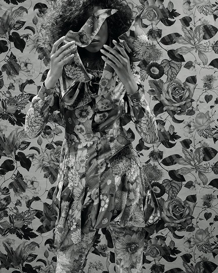 Floral fashion: A person in a floral-print outfit stands against a matching floral background, obscuring their face with floral fabric.