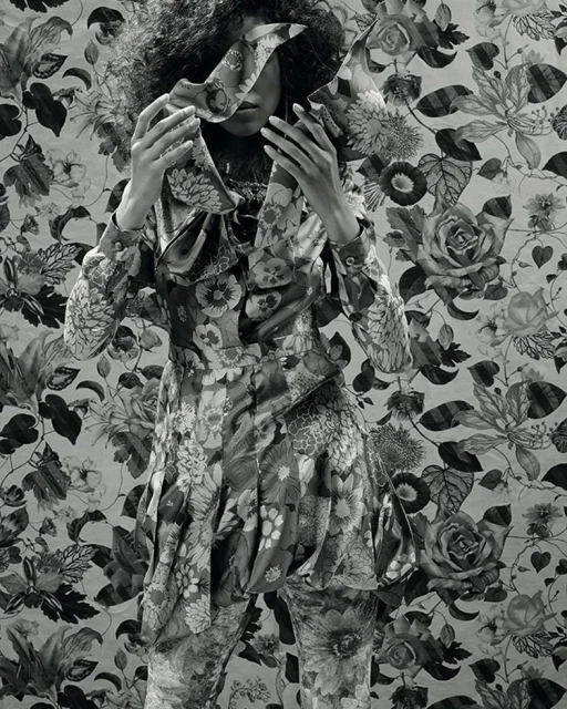 Floral fashion: A person in a floral-print outfit stands against a matching floral background, obscuring their face with floral fabric.