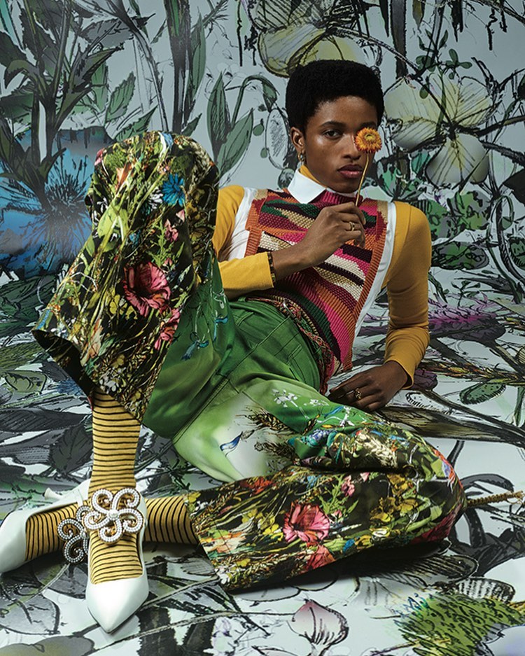 A person poses in colorful, patterned clothing with a flower in front of a painterly floral backdrop in a studio-lit fashion shot.
