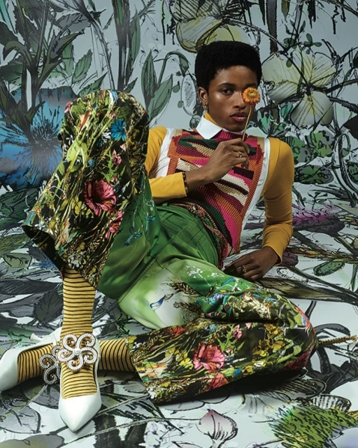 A person poses in colorful, patterned clothing with a flower in front of a painterly floral backdrop in a studio-lit fashion shot.