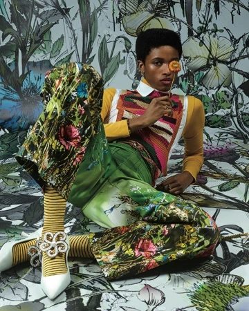 A person poses in colorful, patterned clothing with a flower in front of a painterly floral backdrop in a studio-lit fashion shot.
