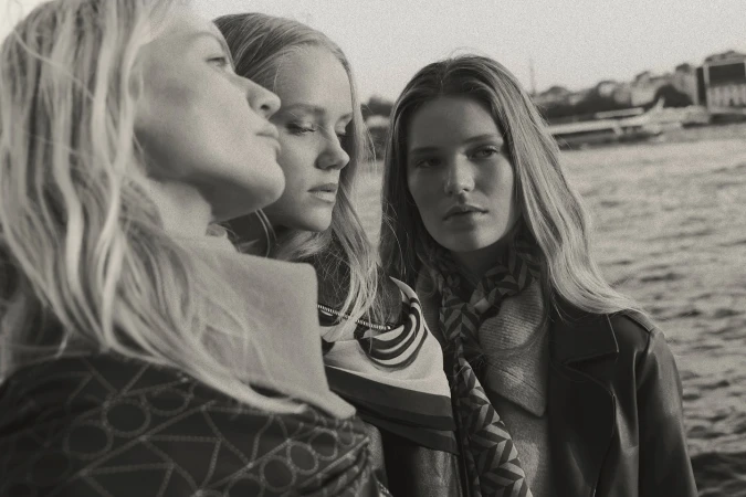 Monochrome portrait of three blonde female models outdoors by the water wearing patterned scarves and coats by Jonas Bresnan