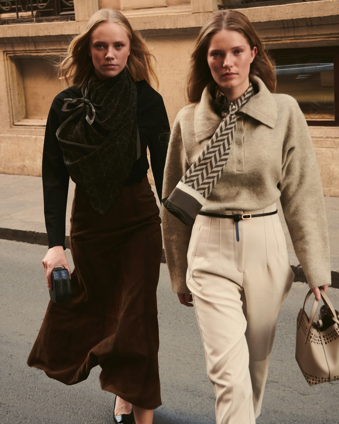Two fair-skinned women walk outdoors in stylish sweaters, scarves, midi skirt, and trousers, carrying handbags against an urban backdrop.