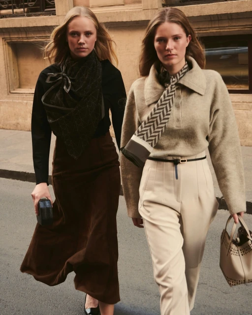 Two fair-skinned women walk outdoors in stylish sweaters, scarves, midi skirt, and trousers, carrying handbags against an urban backdrop.