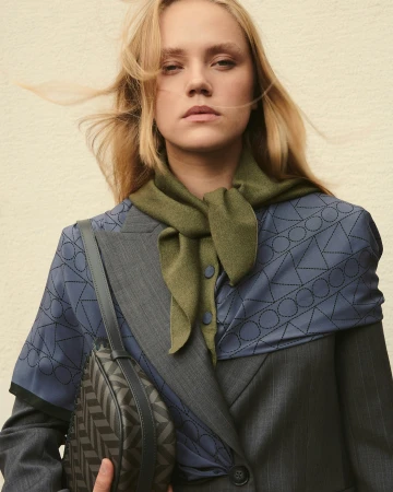 Portrait of a blonde woman wearing an olive green scarf, blue patterned blouse, grey blazer, and holding a patterned shoulder bag.