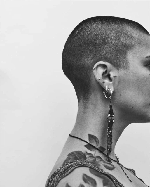 Monochrome close-up of a woman with a shaved head, multiple ear piercings, a rose tattoo on her shoulder, and a long, ornate earring.