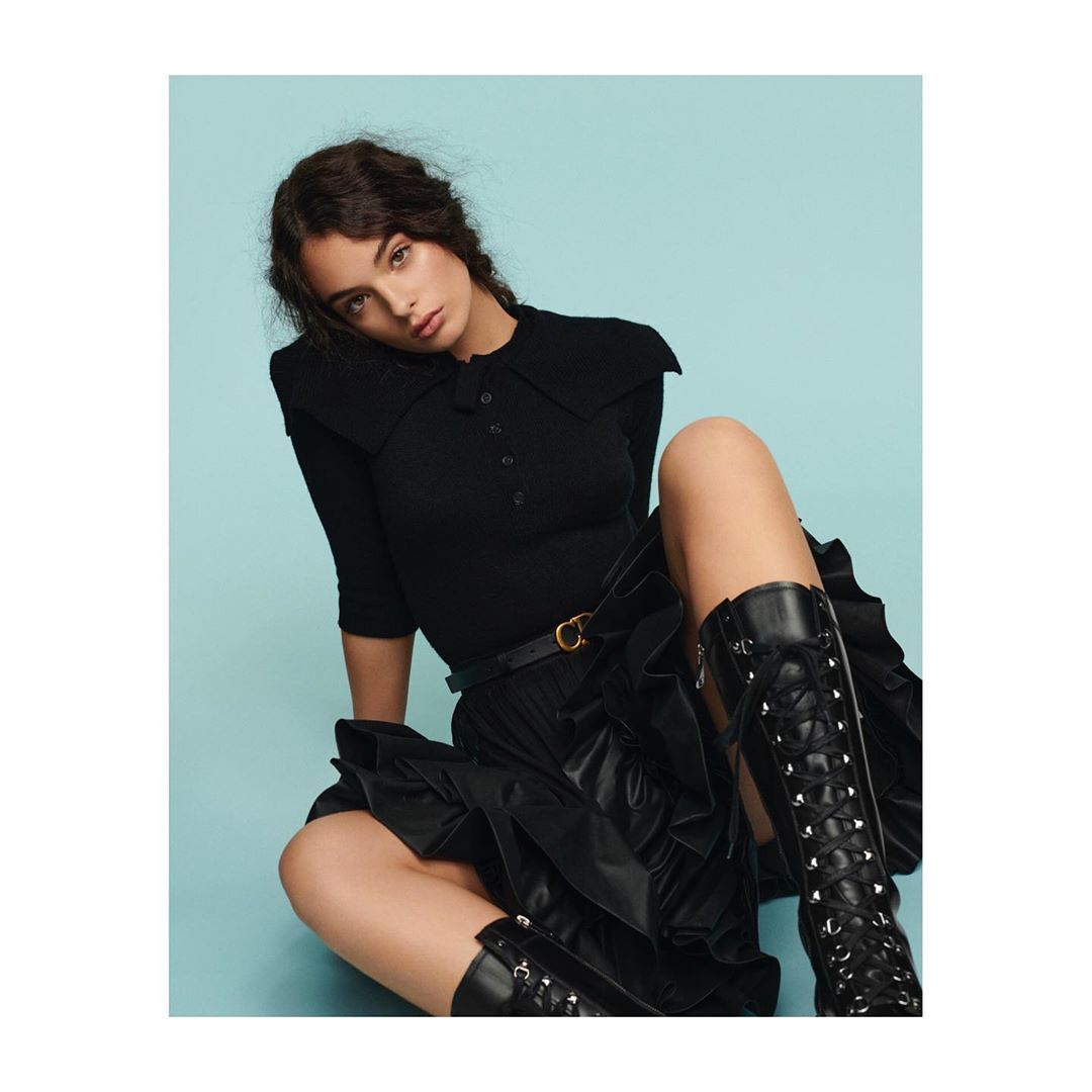 Fashion shot of a female model wearing a black polo top, leather skirt, and lace-up boots against a light blue background.
