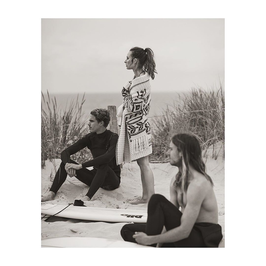 Three people, two men and one woman, pose on a beach; the woman stands wearing a patterned sweater, the men sit nearby.