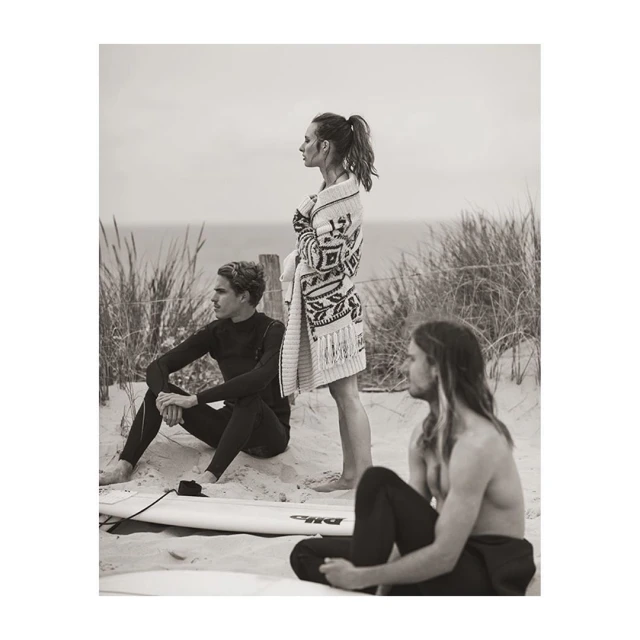 Three people, two men and one woman, pose on a beach; the woman stands wearing a patterned sweater, the men sit nearby.