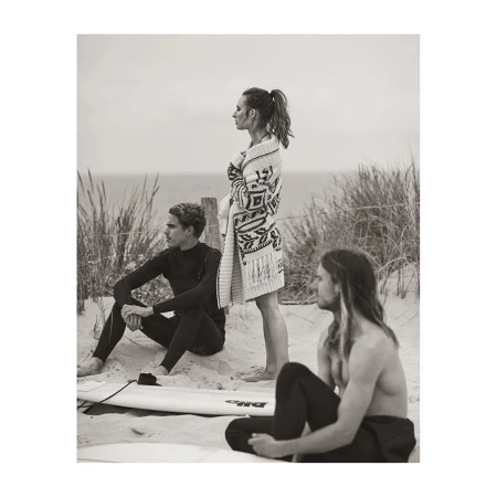 Three people, two men and one woman, pose on a beach; the woman stands wearing a patterned sweater, the men sit nearby.