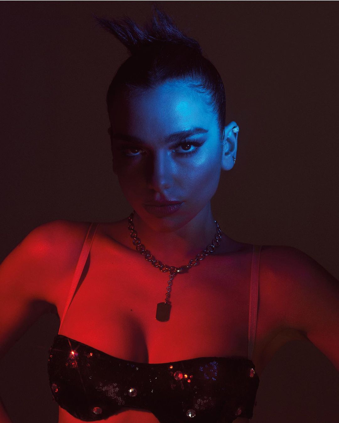 Portrait of a woman with dark hair and strong makeup looking at the camera, set against a dark backdrop with dramatic red and blue lighting.