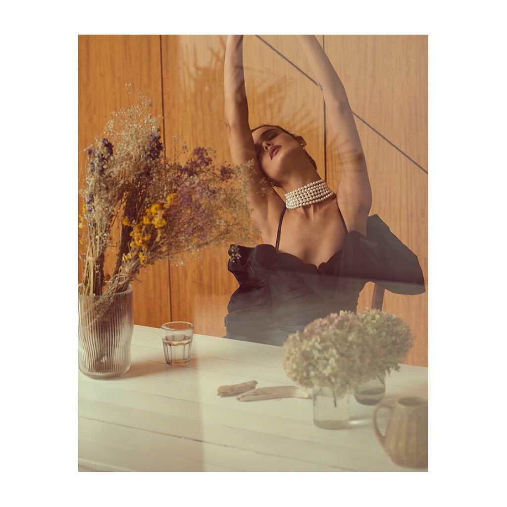 Elegant woman in black dress and pearl necklace sits at a table with floral arrangements in a sunlit room, creating a sophisticated scene.