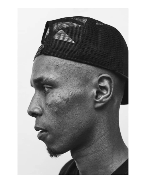 Portrait of a Black adult male in profile wearing a black baseball cap with white background in black and white photography.