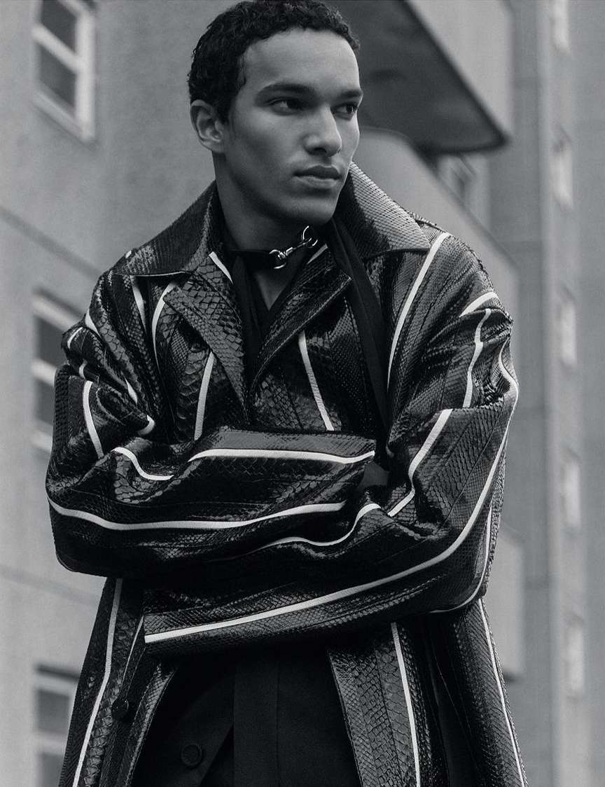 A young adult male poses outdoors against a building while wearing a black snake print trench coat with white stripe details.