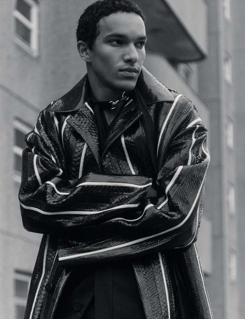 A young adult male poses outdoors against a building while wearing a black snake print trench coat with white stripe details.