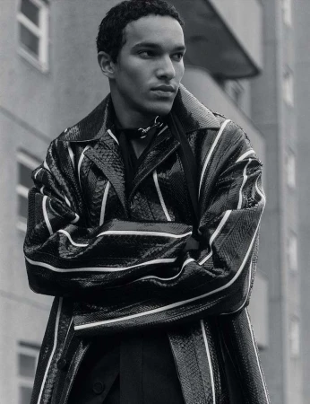 A young adult male poses outdoors against a building while wearing a black snake print trench coat with white stripe details.