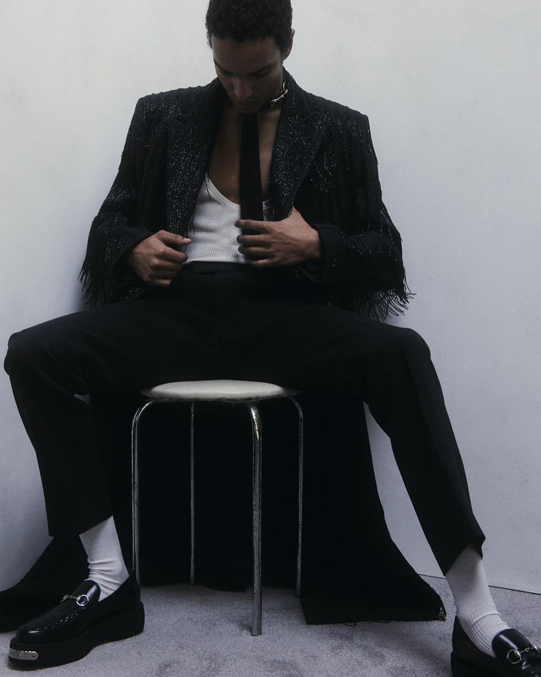 Man seated on a stool wearing a black fringe jacket, white tank top, black tie, dark trousers, and chunky loafers.