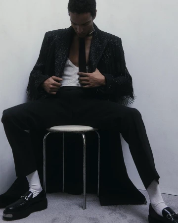 Man seated on a stool wearing a black fringe jacket, white tank top, black tie, dark trousers, and chunky loafers.