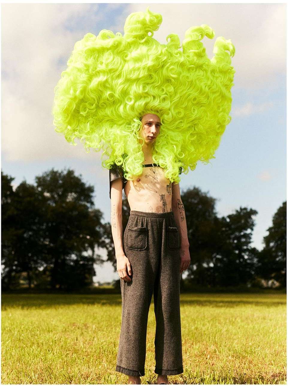 A fair-skinned person with a large, curly, bright-green wig, grey pants and minimal shirt standing in a grassy field with trees in the distance.