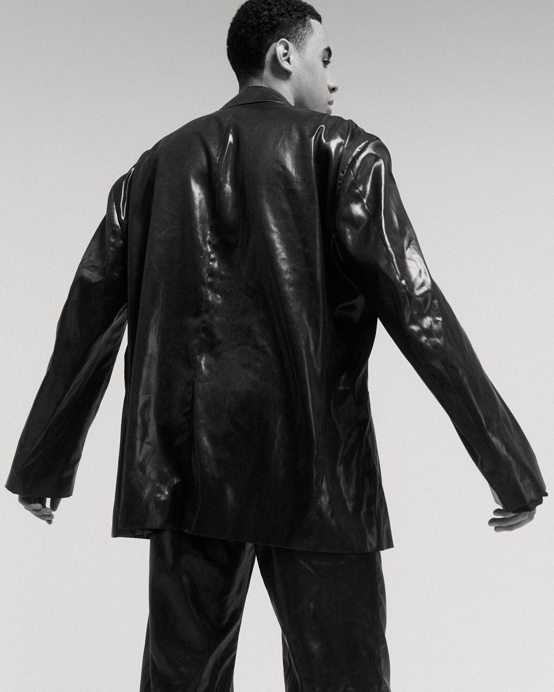 Back view of a Black male model posing in a shiny black suit against a neutral background in a studio setting.