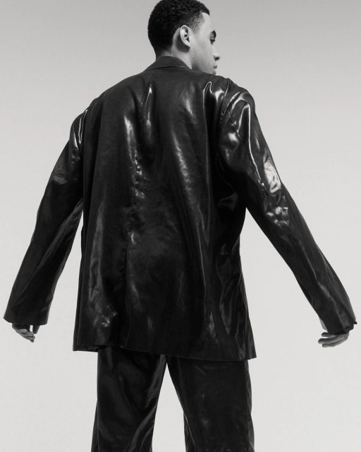 Back view of a Black male model posing in a shiny black suit against a neutral background in a studio setting.