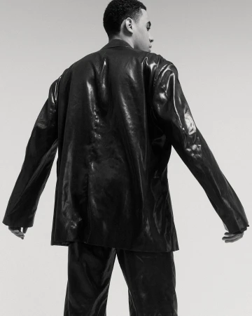 Back view of a Black male model posing in a shiny black suit against a neutral background in a studio setting.