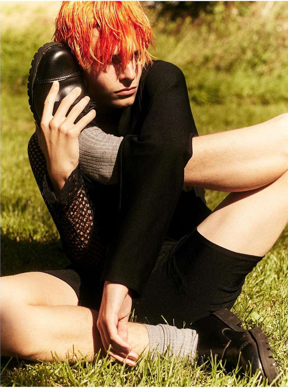 Young adult poses on grassy field with bright orange hair, wearing a black top and shorts, hugging a black shoe, editorial fashion.