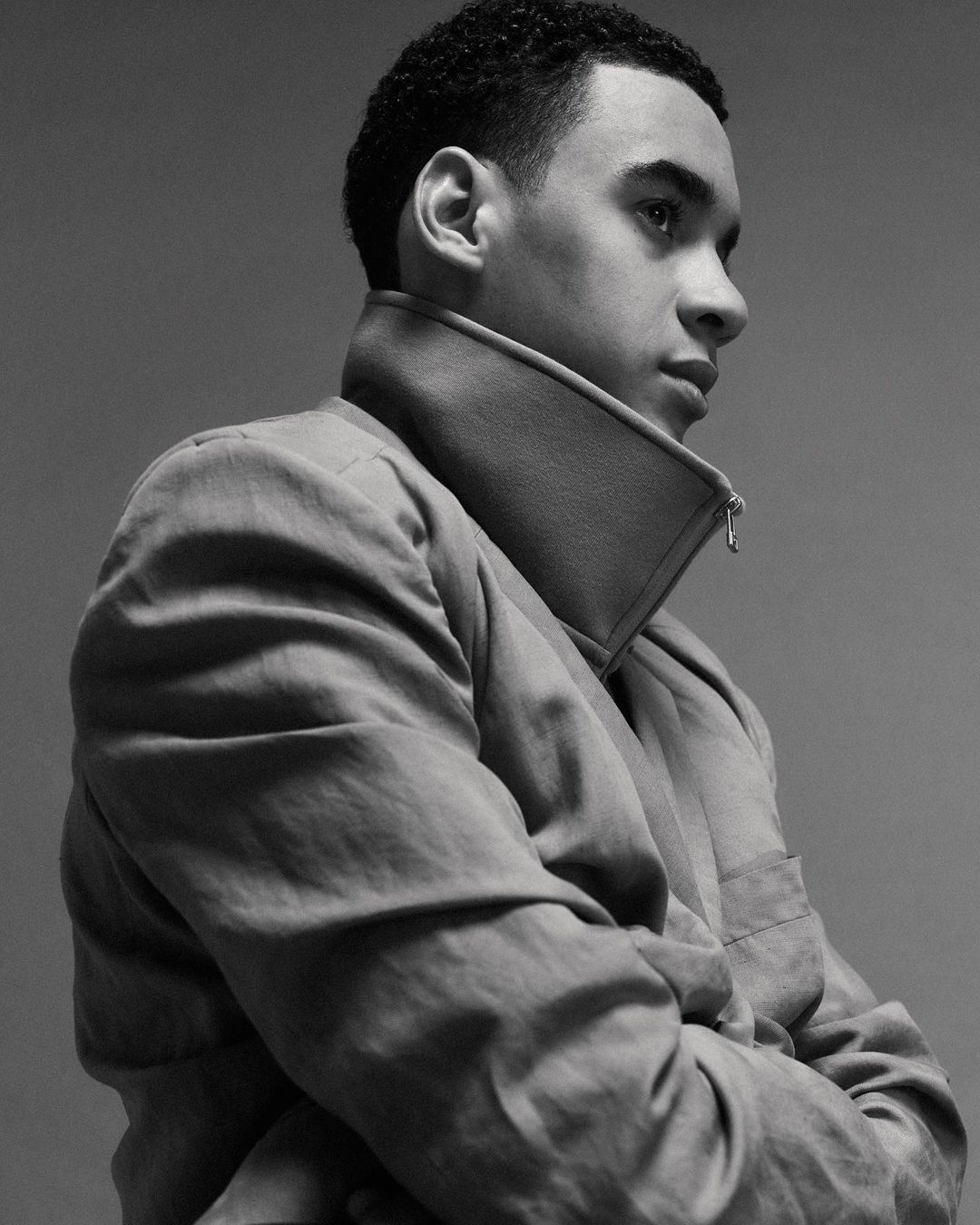 Black and white portrait of a young adult male with short curly hair in a light-colored jacket, posing with arms crossed in a studio.