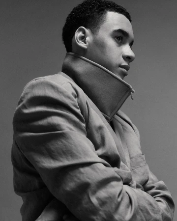 Black and white portrait of a young adult male with short curly hair in a light-colored jacket, posing with arms crossed in a studio.