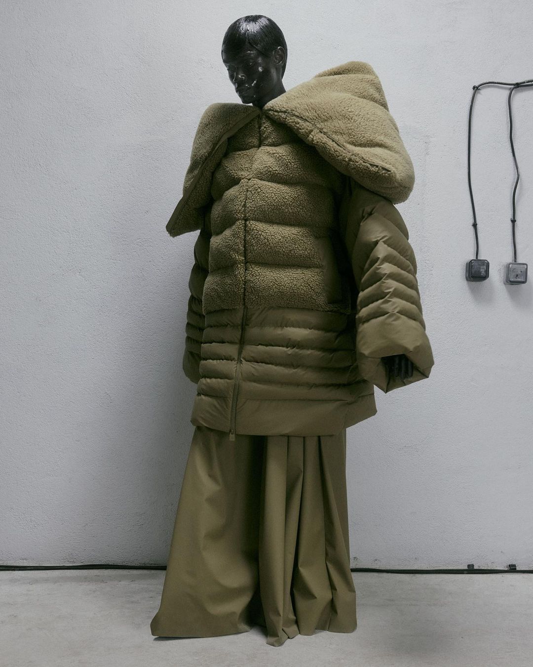 Mannequin displays a puffy olive-green coat with a large hood, paired with a long skirt in a minimalist indoor setting.