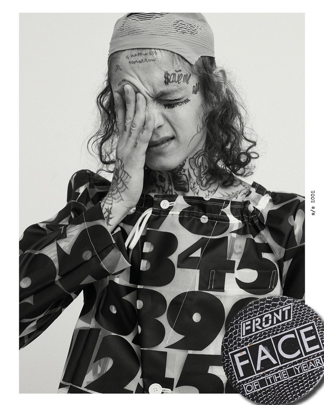 Black and white portrait of a young adult with face tattoos, hand on face, wearing a printed shirt, and a headscarf.