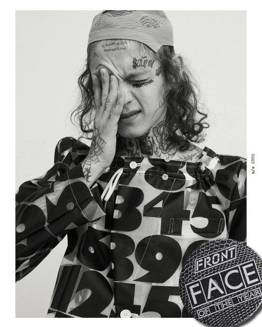 Black and white portrait of a young adult with face tattoos, hand on face, wearing a printed shirt, and a headscarf.