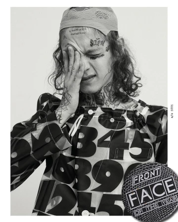 Black and white portrait of a young adult with face tattoos, hand on face, wearing a printed shirt, and a headscarf.