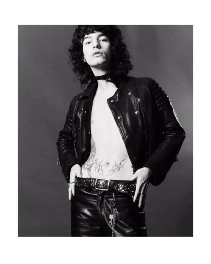 Monochrome studio portrait of a young adult with curly hair wearing a black leather jacket, studded belt, and leather pants posing.