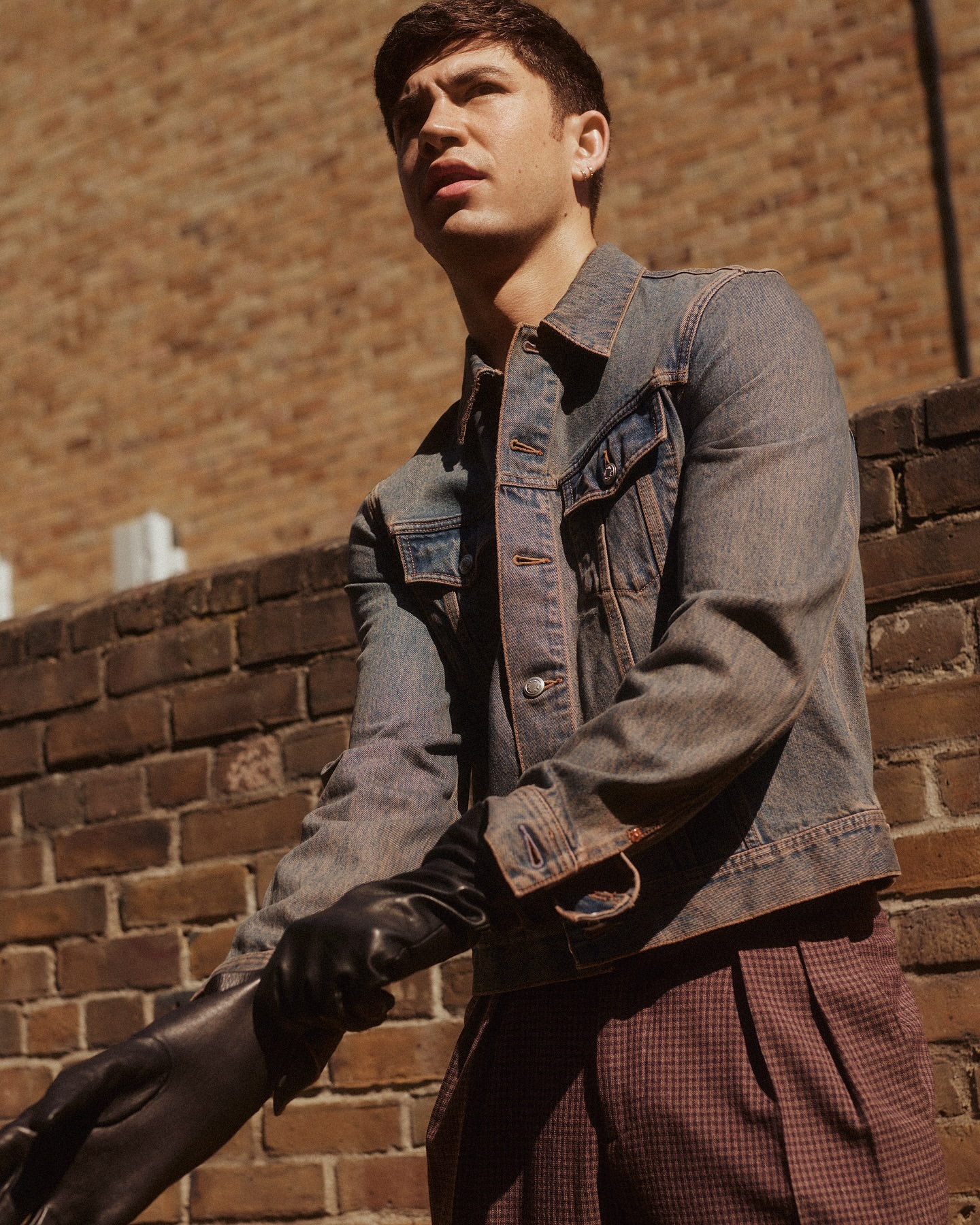Young man poses outdoors against a brick wall, wearing a denim jacket, plaid pants, and black gloves, glancing to the side in soft light.