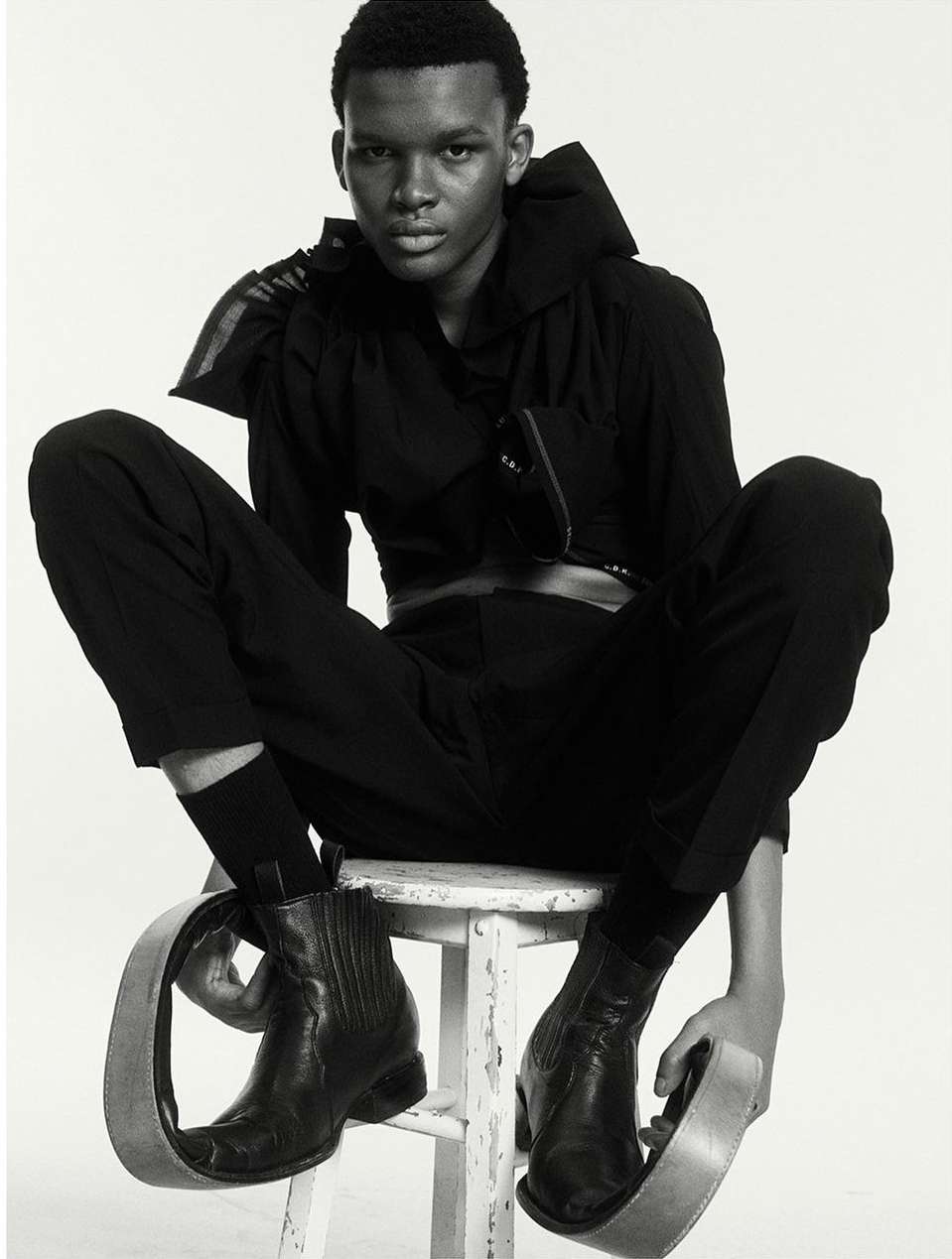 An androgynous-looking person sits on a white stool wearing a black outfit, cowboy boots, and metal loop accessories on their feet.