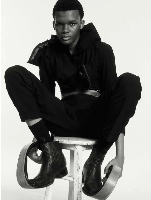 An androgynous-looking person sits on a white stool wearing a black outfit, cowboy boots, and metal loop accessories on their feet.