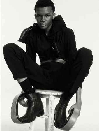 An androgynous-looking person sits on a white stool wearing a black outfit, cowboy boots, and metal loop accessories on their feet.