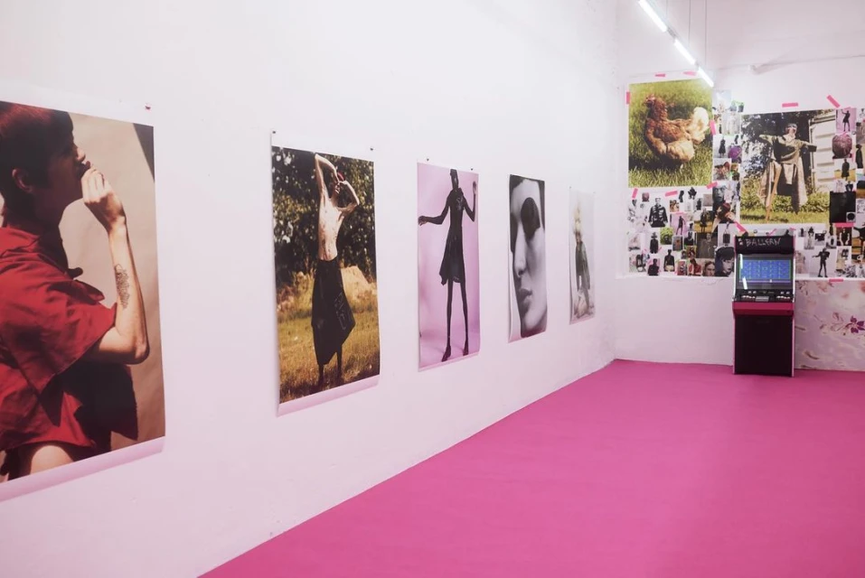 Art gallery view with large format photographs on white walls, pink floor, and a vintage arcade game in the corner of the room.