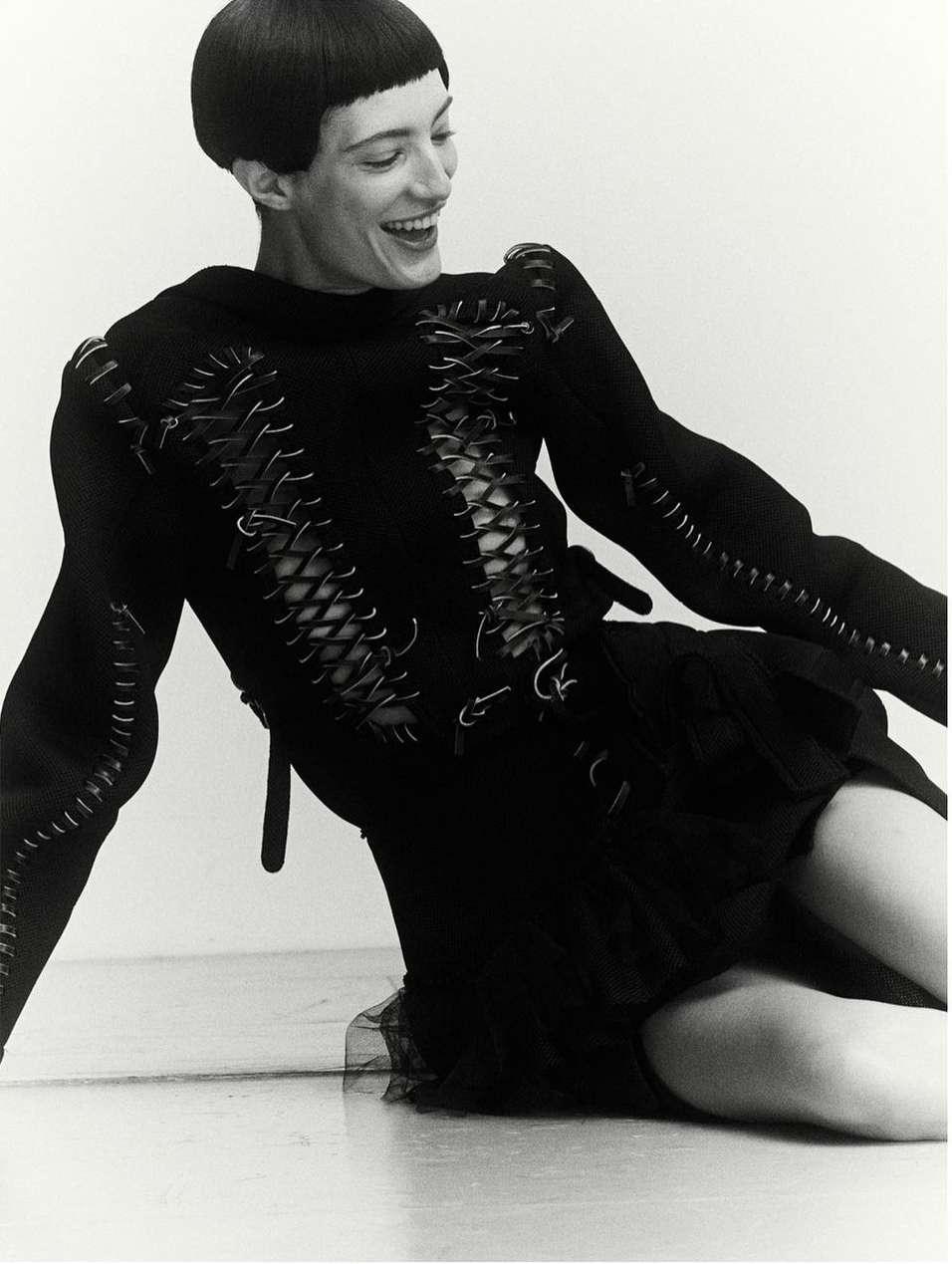 A smiling woman with short dark hair is kneeling in a studio setting. She wears a black dress with decorative stitching details and corset-style lacing.