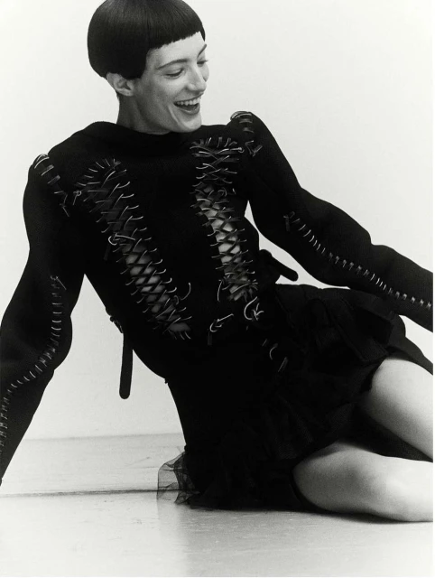A smiling woman with short dark hair is kneeling in a studio setting. She wears a black dress with decorative stitching details and corset-style lacing.