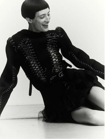 A smiling woman with short dark hair is kneeling in a studio setting. She wears a black dress with decorative stitching details and corset-style lacing.