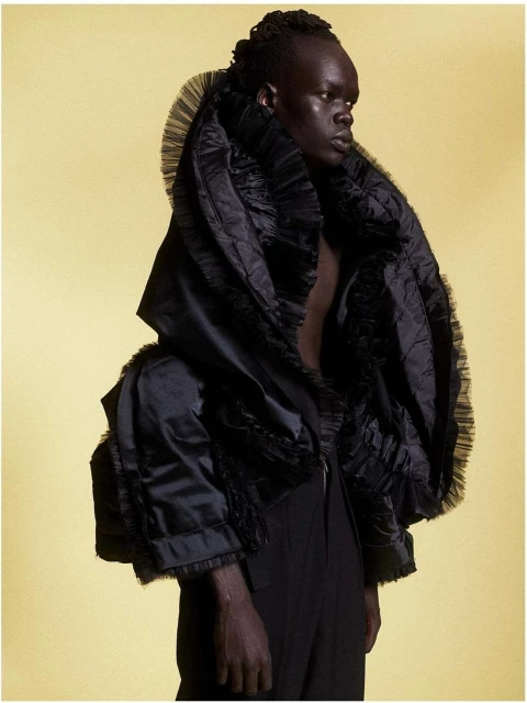 Portrait of a person with dark skin wearing a voluminous black ruffled garment against a pale yellow backdrop in a studio setting.
