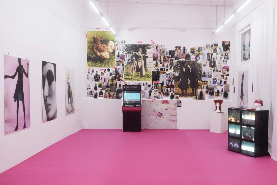 Art gallery exhibition space with posters and images on white walls and pink floor, featuring vintage electronics and display monitors.